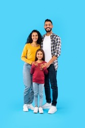 Cheerful Arabic Family Of Three Posing Together, Father And Mother Embracing Their Kid Daughter Standing On Blue Studio Background. Full Length, Vertical Shot