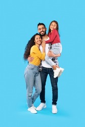 Arabic Man Hugging Wife And Holding Little Daughter In Arms Standing On Blue Background. Happy Middle Eastern Family Posing In Studio Smiling To Camera. Vertical Shot