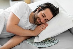 Smiling bearded young guy sleeping with bunch of cash under his pillow, keeping his money savings by him at home, closeup photo. Finance management concept