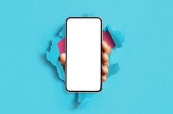 Mobile App, Great Offer. Black male hand holding smartphone with white empty screen showing device close up to camera breaking through blue paper sheet. Gadget display with free copy space, mock up