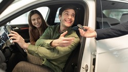 Happy millennial couple taking car key from auto salesman, sitting inside modern automobile at dealership, panorama. Cheery young family buying new vehicle at modern showroom