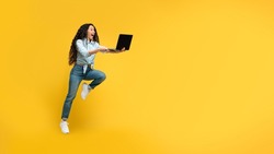 Full body length of funky woman jumping high in the air holding and using pc laptop, browsing social media online, searching discounts and sales, yellow orange wall, banner panorama, free copy space