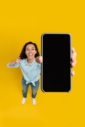 Cheerful young woman holding cell phone in hand pointing at gadget with blank black mockup screen smiling on yellow orange wall, recommending newest mobile app, above high angle top view, copy space