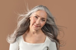 Portrait of smiling senior woman posing with long and healthy grey blowing flying hair on brown studio background. Beautiful mature lady posing and smiling at camera. Haircare and fashion concept