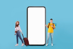 Cool Mobile Offer For Travellers. Couple of tourists presenting big giant cell, excited guy with backpack pointing at huge white blank screen, lady holding bag on blue studio wall, full body length