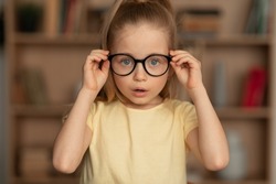 Little Girl Wearing Eyeglasses Having Poor Eyesight Posing Looking At Camera Through Eyewear Standing Near Bookshelf At Home. Kid's Eyes Health. Knowledge And Reading Concept