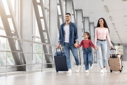 Happy Middle Eastern Family Of Three Walking With Suitcases At Airport Hallway, Cheerful Arabic Parents And Little Daughter Travelling Abroad Together, Holding Hands And Smiling, Copy Space