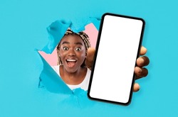 Glad excited surprised young african american lady with open mouth looks through hole in blue paper and shows smartphone with blank screen, studio. Online win, sale, great offer, app and new device