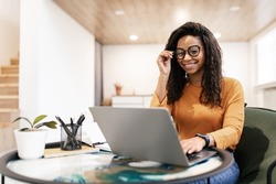 People And Technology. Portrait of young black woman wearing eyeglasses using pc sitting at table on beanbag chair in living room, typing on keyboard. Cheerful lady browsing internet, free copy space