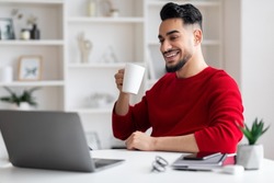 Happy young islamic man with beard drinks cup of tea at workplace with computer in home office interior. Business, coffee break from work, pause and vitality for freelance during covid-19 outbreak