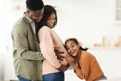 Joy Of Pregnancy. Happy Black Family Hugging Expecting Baby, Daughter Listening To Pregnant Mommy's Belly Standing In Modern Kitchen At Home. Childbirth And Parenting Concept