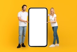 Happy young caucasian male and female in white t-shirts presentation huge phone with empty screen isolated on yellow background, studio, full length. Emotions from ad and sale, great offer and app