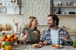 Laughing happy european millennial blond wife and hasband with stubble eating pasta in light kitchen interior. Romantic dinner at home together, love, relationship and family during covid-19 outbreak