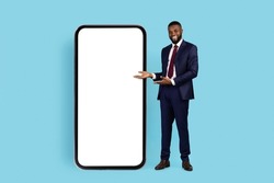 Successful Black Businessman Pointing At Big Smartphone With White Blank Screen, African American Male In Suit Showing Free Copy Space For Mobile Business App Or Website Design, Mockup
