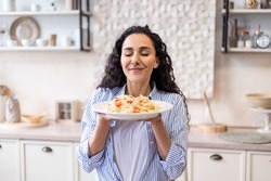 Happy lady tasting homemade spaghetti while having lunch, enjoying the smell with eyes closed, sitting in kitchen interior. Woman cooking and eating tasty food at home