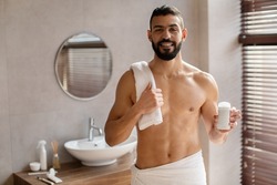 Advertisement Concept. Happy shirtless guy holding tube of deodorant in hand standing in modern bathroom. Smiling man presenting tube of conditioner and lotion, showing cosmetic product, stick package