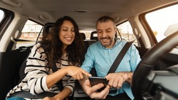 Short Road Route. Portrait of smiling couple sitting inside luxury car pointing fingers at smart phone, cheerful male driver holding cell, showing choosing location, using digital map application