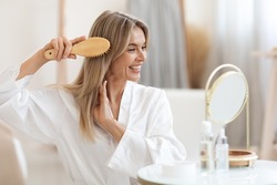 Smiling beautiful middle aged blonde woman in white silky bathrobe sitting in front of mirror at bathroom, combing her smooth hair with wooden brush at home, copy space