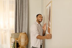 Smiling young black man putting picture frame, hanging painting on wall, empty space. African American male interior designer decorating new modern stylish apartment. Home interior and domestic decor