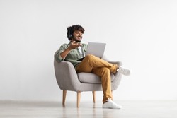 Positive handsome young indian guy sitting in armchair over white studio background, using laptop and headset, having video call with friend or lover, copy space. Modern technologies and communication