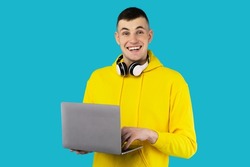 Happy Student Guy Using Laptop Computer Posing With Wireless Headphones Smiling To Camera Standing In Studio Over Blue Background, Wearing Yellow Hoodie. E-Learning And Technology