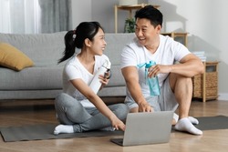 Cheerful asian lovers in sportswear sitting on fitness mat in front of computer, resting after online yoga class, having conversation, drinking water, home interior, copy space