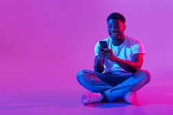 Full length of millennial African American guy sitting cross legged with smartphone, surfing social media, chatting to friend in neon light, copy space. Cool black man using mobile phone