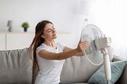 Glad young european woman suffers from heat sits on sofa catches cold air, turns fan to herself, enjoys cool down in living room interior. Summer weather, overheating without air conditioning at home