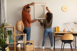 Black Millennial Spouses Hanging Poster In Frame On Wall Together Standing At Home. Married Couple Decorating Living Room After Renovation In New House Indoors. Interior Decoration And Design
