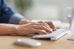 Unrecognizable male employee working on laptop at desk in office, cropped image of young man typing on computer keyboard, writing email or communicating online, side view shot with selective focus