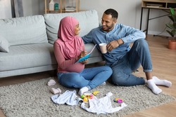 Pregnant black muslim couple making checklist of necessities while getting ready for childbirth, happy islamic spouses awaiting baby, relaxing on floor in living room with child clothes around