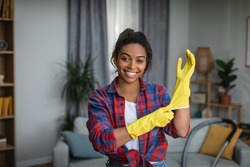 Smiling young african american woman puts on rubber gloves and ready to homework in living room interior. Housewife has fun and many household chores, domestic work and professional cleaning service