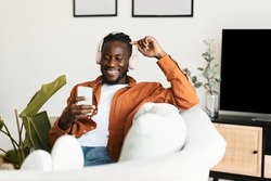 Excited black guy in headphones enjoying music on smartphone and scrolling social networks feed, resting on sofa at home, free space. Happy man relaxing to favorite melody indoors
