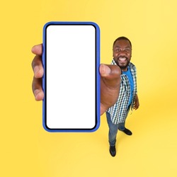 African American Male Holding Huge Mobile Phone Showing Blank Screen To Camera Advertising Application Standing On Yellow Studio Background. High Angle Shot, Square, Mockup