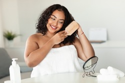 Happy african american oversize woman brushing her curly hair with comb while sitting near mirror in bedroom. Positive black lady wrapped in towel making haircare routine