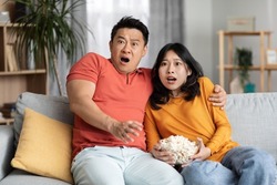 Thrilled korean man and woman watching spooky movie at home, middle aged husband hugging his young wife, sitting on couch, eating popcorn, spending time together, copy space