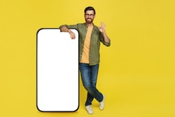 Positive stylish man showing okay gesture, standing next to huge smartphone with blank screen over yellow background, space for mobile app or website design on screen. Advertisement mockup