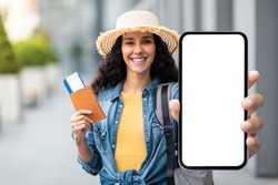 Happy attractive millennial brunette lady in straw hat tourist holding passport with flight tickets and cell phone with white blank screen, showing nice mobile app for travellers, mockup