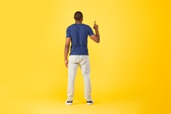 Back View Of African American Man Pointing Finger Up Pushing Invisible Button Or Touchscreen Standing Over Yellow Studio Background. Look Upward. Full Length Shot