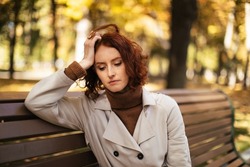 Sad tired young european lady in raincoat sits on bench, thinks, suffer from headache and migraine in park in autumn. Emotions, health care problems, stress and pressure during covid-19 quarantine