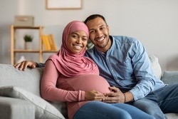 Portrait Of Happy Black Muslim Couple Expecting Baby Posing In Home Interior, Joyful Young Islamic Spouses Sitting On Couch In Living Room And Smiling At Camera, Husband Touching Wife's Belly