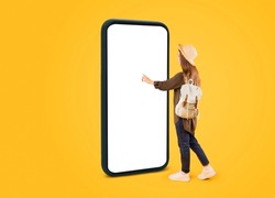 Vacation Offer. Tourist Woman Using Huge Cellphone With Empty Touchscreen Booking Travel Tickets Online Standing In Studio Over Yellow Background. Touristic App. Mockup