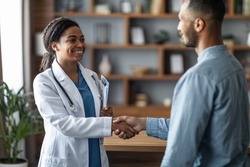 Cheerful african american young woman doctor greeting patient handsome millennial middle eastern man, doc and patient shaking hands and smiling, clinic interior, copy space