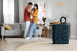 Selective focus on brand new portable speaker over dancing romantic asian couple, blurred background, copy space, home interior. Modern technologies and domestic entertainment concept