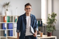 Portrait Of Handsome Young Male Entrepreneur Holding Laptop And Posing In Office Interior, Confident Millennial Man In Casual Clothes Standing Near Desk And Smiling At Camera, Copy Space