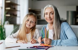 Cheerful caucasian little girl with aged lady with colorful pencils draw a picture on table, enjoy study and free time in living room interior. Art lesson together, education and entertainment at home