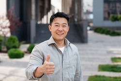 Portrait of positive asian man standing outdoors and gesturing thumb up, walking in urban city area, looking and smiling at camera, free copy space