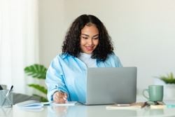 Black Businesswoman Using Laptop Computer Taking Notes Sitting In Modern Office Indoors. Successful Business Lady Working Online At Workplace. Female Entrepreneurship Career Concept