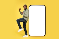 Glad mature african american male in casual rejoices to victory near huge smartphone with empty screen, isolated on yellow background, studio. Celebrate success and online win, new app, ad and offer