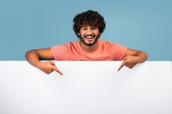 Smiling young hindu man with curly hair in casual outfit pointing down at white blank advertising board with empty space for text, blue studio background, mockup, panorama with copy space